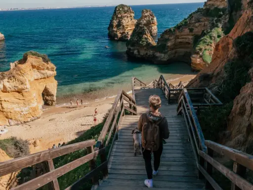 Persona desciende escaleras de madera hacia una hermosa playa de lagos con formaciones rocosas y aguas turquesas.