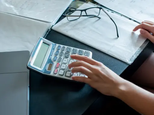 Ręce liczą na kalkulatorze dokumenty, analizując umowę zlecenie a alimenty. Obok leżą okulary i notatki.