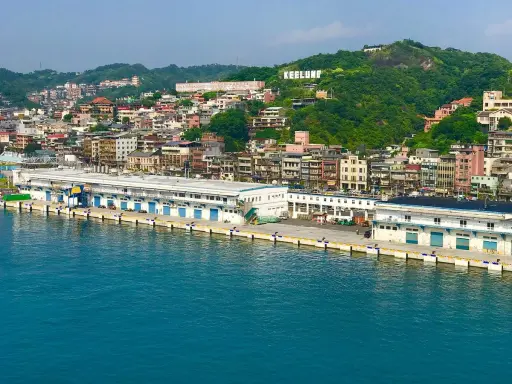 Vista panoramica del porto di Taiwan, con edifici colorati che si arrampicano sulla collina verde. La scritta "KEELUNG" domina la cima.