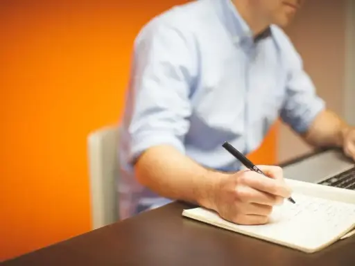 Mężczyzna w niebieskiej koszuli pisze piórem w notatniku, obok laptopa. Działa z pełną **prokura**.