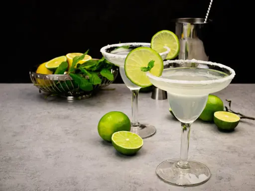 Zwei Margaritas mit Salzrand, garniert mit Limettenscheiben und Minze. Limetten und Minze in einer Schale im Hintergrund. Perfekt für ein Margarita Rezept.