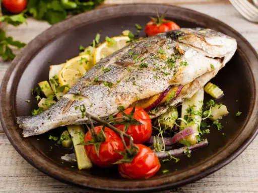 Poisson entier, probablement une truite au barbecue, servi avec des tomates cerises, des quartiers de citron et des herbes fraîches sur une assiette marron.