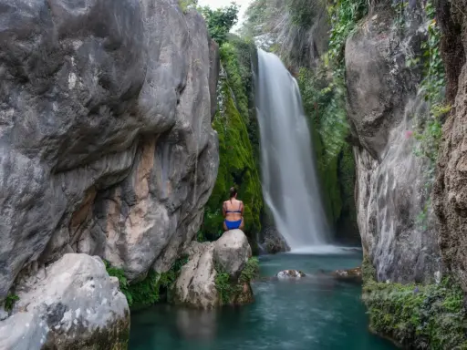Una mujer sentada en una roca contempla la cascada de las fuentes del Algar, rodeada de vegetación y rocas.