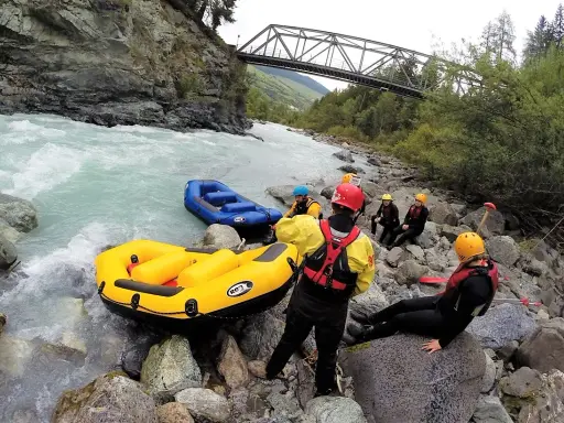 Grupa w kamizelkach ratunkowych i kaskach przygotowuje się do raftingu na rwących rzekach w Austrii. Dwie dmuchane łodzie czekają na brzegu.