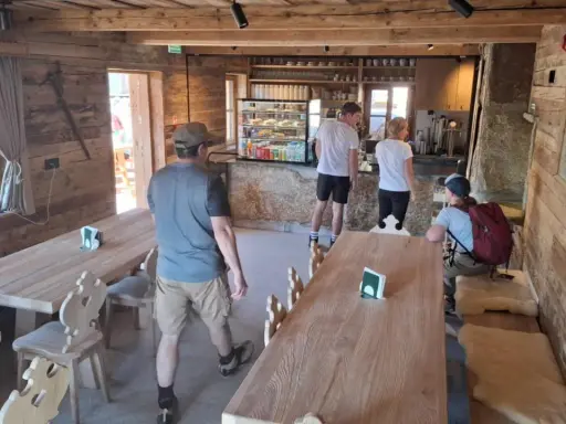 Wnętrze schroniska na Hali Kondratowej. Drewniane stoły i ławy, bar z ekspozycją ciast i napojów. Kilka osób odpoczywa po wędrówce.