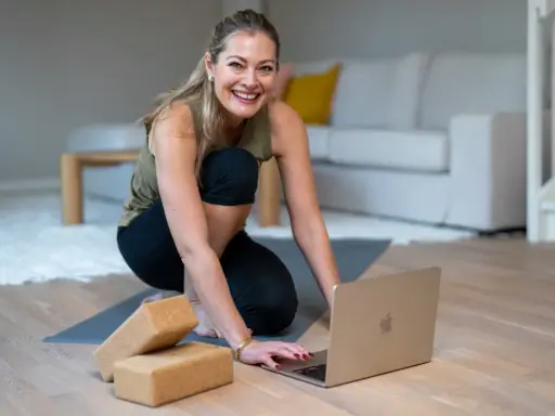Glad kvinna förbereder sig för en lugn stund med **restorative yoga** hemma, med en laptop och yogablock redo.