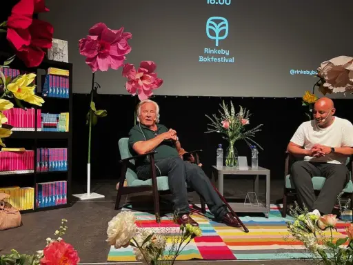 Jan Guillou delar fakta på Rinkeby Bokfestival. Scenen är dekorerad med stora blommor och bokhyllor.