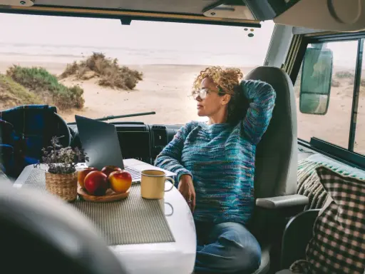 Donna con capelli ricci lavora al computer in un camper, godendosi la vista del mare. Un esempio di nomade digitale lavoro.