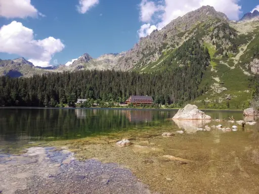Spokojne Popradske Pleso odbija góry i las. Kamienie w płytkiej wodzie tworzą ścieżkę.