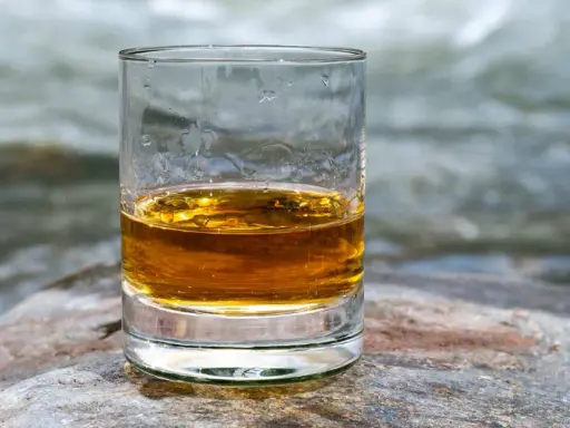 Ein Glas goldener Whisky steht auf einem Stein am Flussufer. Die ideale trinktemperatur für diesen edlen Tropfen.