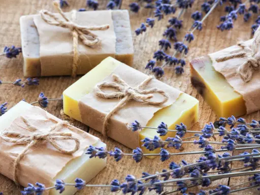 Quattro saponette fatte in casa, avvolte in carta e legate con spago, giacciono su un tavolo di legno rustico, circondate da rametti di lavanda profumata.