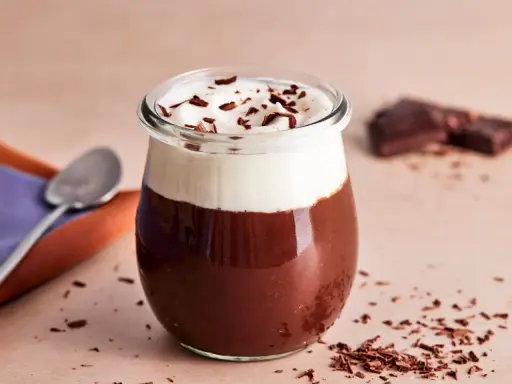 Ein Glas mit Omas Schokoladenpudding mit Sahne und Schokostreuseln. Daneben liegen ein Löffel und ein Stück Schokolade.