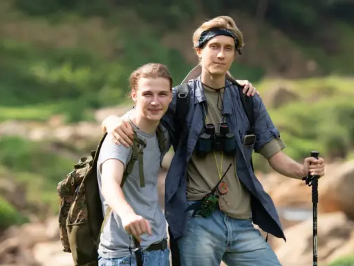 Dwóch młodych mężczyzn, gotowych na trekking, z plecakami i kijkami, w otoczeniu zieleni i skał.