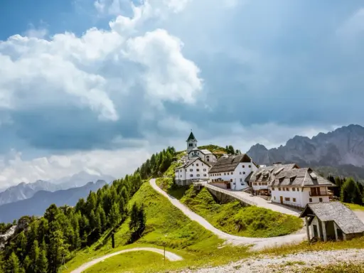 Un borgo incantevole in montagna, perfetto per le tue vacanze in Friuli. Case bianche, una chiesa e sentieri verdi sotto un cielo nuvoloso.