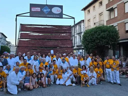 Grupo posa en la fiesta longaniza Graus, con un cartel de "Los Tres Sabores de la Longaniza de Graus" y una estructura de madera con embutidos.