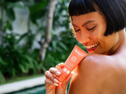 Une femme souriante applique une crème solaire SPF 50+ sur son épaule. Le tube indique "Très haute protection", le SPF 50 signification est une protection optimale.