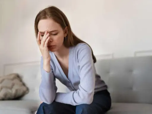 En kvinna sitter på en soffa med handen mot pannan, uttrycker smärta och utmattning i akut fas.
