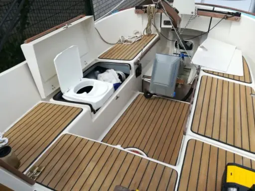 Ein weißes **Segelboot Toilette** mit offenem Deckel, daneben ein grauer Tank. Holzdecks und weiße Wände.