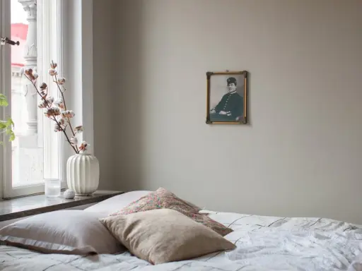 En säng med flera kuddar i olika nyanser av beige och mönster. En gammal porträttbild hänger på väggen.