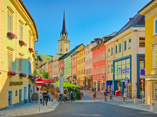 Via colorata di una città austriaca più vicina all'Italia, con edifici vivaci, una torre dell'orologio e gente che passeggia.
