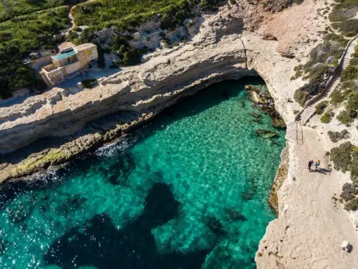 Malta, gdzie na malta? Widok z lotu ptaka na turkusową zatokę, klify i mały domek. Dwie osoby spacerują ścieżką.