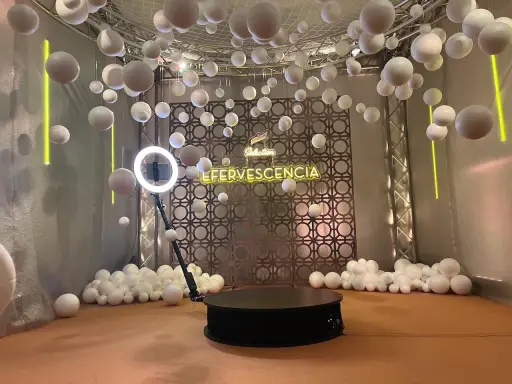 Plataforma giratoria para fotos con luces de neón "EFERVESCENCIA" y esferas blancas colgantes, creando un efecto de bullet time.