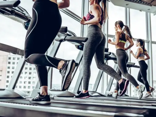 Kobiety ćwiczą cardio na bieżni, biegnąc w siłowni z widokiem na miasto.