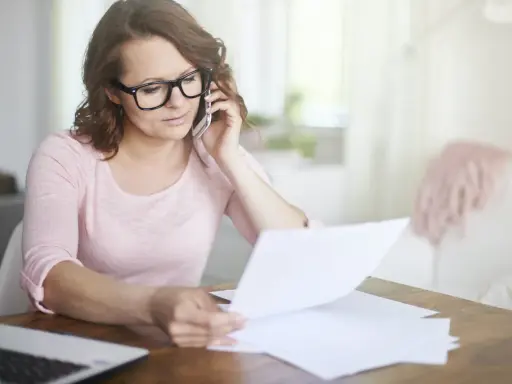 Kobieta w okularach rozmawia przez telefon, przeglądając dokumenty. Martwi się, że czeka ją brak świadectwa pracy.