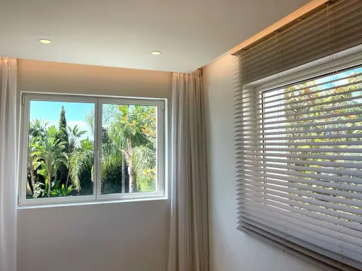 Dos tipos de apertura de ventanas: una corredera con cortinas y otra con persianas venecianas, ambas con vistas a un exuberante jardín tropical.