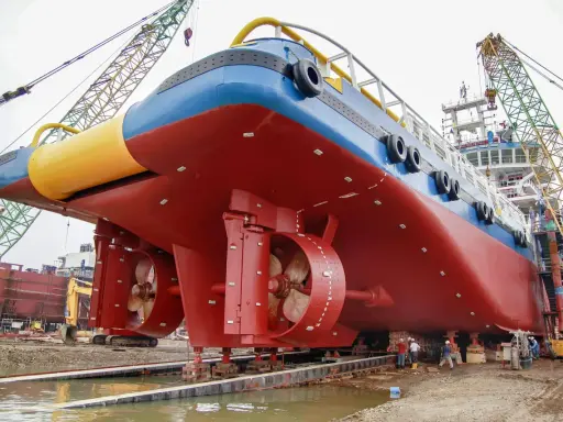 Gros navire rouge et bleu sur des supports, avec des hélices visibles. Des grues et des ouvriers sont présents. On aperçoit des anodes sur la coque, protégeant contre la corrosion, qu'elles soient anode + ou -.