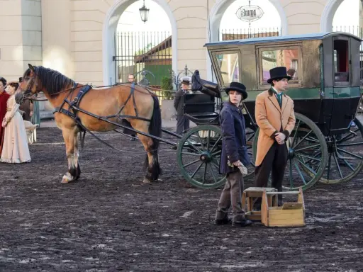 Konny zaprzęg z dorożką i postaciami w strojach z epoki na tle budynków.