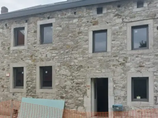 Maison ancienne en cours de rénovation, avec des murs en pierre brute et de nouvelles fenêtres. Le projet de construire en pierre continue.
