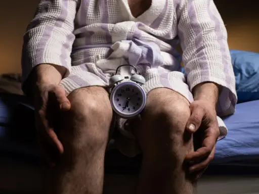 Mężczyzna w szlafroku siedzi na łóżku z budzikiem między kolanami. Czas na rozmowę o andropauzie a seksualności.