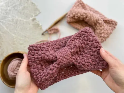 Manos sostienen una diadema tejida a crochet con un lazo. Detrás, otra diadema y ovillo de lana. ¡Descubre patrones gratis de diademas de ganchillo!