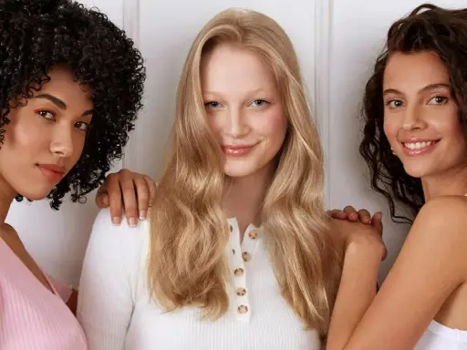 Drei Frauen stehen nebeneinander. Die Frau in der Mitte hat orange blonde haare und lächelt.