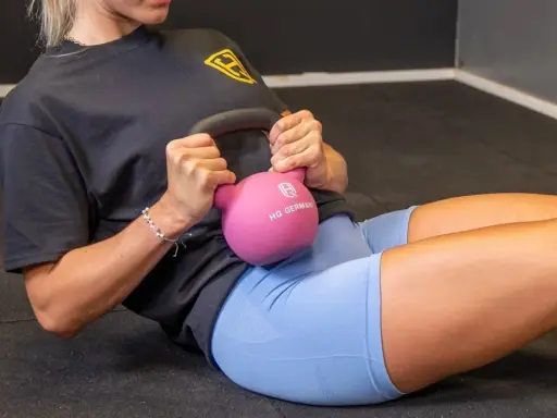 Frau macht ein **Kugelhantel Training** auf dem Boden, hält eine pinke Kettlebell und trainiert ihre Bauchmuskeln.