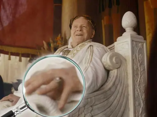Ein Mann mit Lorbeerkranz, der an alle Kaiser Roms erinnert, sitzt auf einem Thron. Eine Lupe enthüllt Details seiner Hand.
