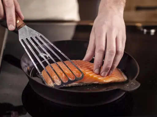 Ein Lachsfilet wird mit einem Pfannenwender in eine heiße Pfanne gelegt. Perfekt für leckere Fisch Rezepte!