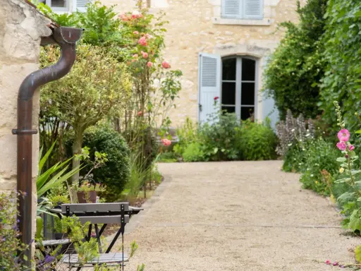 Un chemin de gravier serpente à travers un jardin anglais romantique, bordé de roses roses et de verdure luxuriante. Une maison en pierre avec des volets bleus se profile à l'arrière.