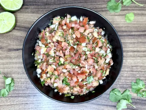 Świeże, kolorowe pico de gallo w czarnej misce, z limonkami i kolendrą na drewnianym tle.