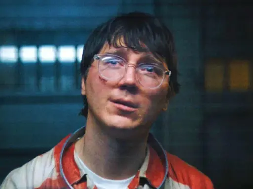 Paul Dano med glasögon och blodfläckar på sin randiga skjorta, i en mörk miljö med ljus bakom.