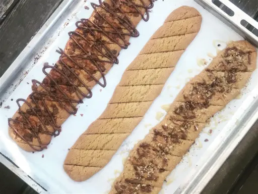 Tre långa, avlånga kakor på en plåt. En är dekorerad med chokladglasyr och hackad choklad, en annan med strössel och en tredje med honung och nötter. Perfekt för ett kolakakor recept!