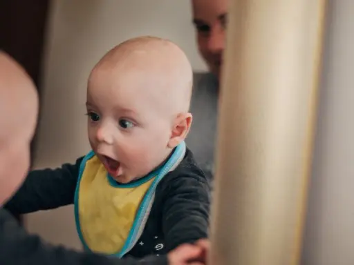 Ein Baby mit gelbem Lätzchen schaut überrascht in einen Spiegel. Vielleicht fragt es sich, ab wann Kinder "Ich" sagen.