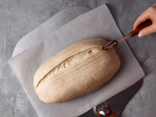 Ein Laib Brot wird mit einem Bäckermesser eingeschnitten, bereit zur Zubereitung mit Sauerteigpulver.