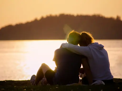 Ein Paar sitzt am Seeufer im Sonnenuntergang, die Arme umeinander geschlungen. Sie finden Trost und lernen, sich selbst zu verzeihen.