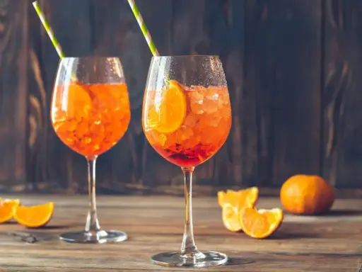 Dwa orzeźwiające Aperol Spritz z plasterkami pomarańczy i słomkami, na drewnianym stole.