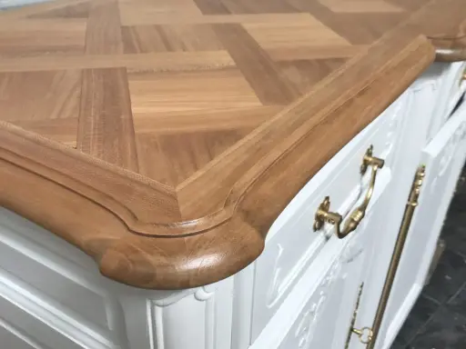 Un meuble ancien relooké en moderne, avec un plateau en bois clair à motif géométrique et des poignées dorées sur un corps blanc.