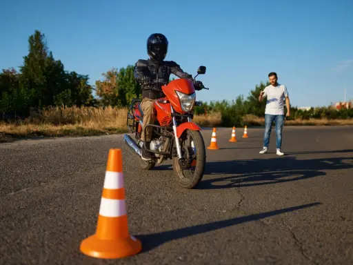 Persona con casco y equipo de protección conduce una moto naranja en un circuito de prácticas. Un instructor observa.