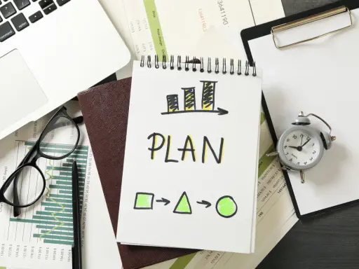Na biurku leżą dokumenty, laptop, okulary i budzik. Na notatniku widnieje napis "PLAN" i schemat, który symbolizuje indywidualny plan rozwoju.