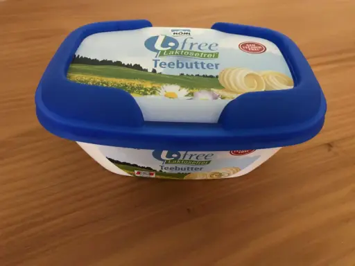 Eine Packung "Biofree Laktosefrei Teebutter" mit blauem Deckel steht auf einem Holztisch. Die Verpackung zeigt eine Sommerwiese mit Blumen und Butterflocken.